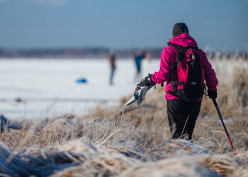 En person i rosa jacka går över frosigt gräs mot en frusen sjö. Hen har långfärdsskridskor och stavar i handen.
