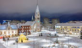 Vy över ett snötäckt Hertig Johans torg med kyrkan i bakgrunden och stämningsfull julbelysning. 