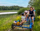 Two blue railbikes with families on them. They are cycling in a green landscape by the water.