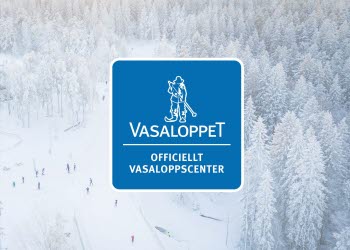 Skidåkare i skidspåret genom en snötäckt skog. Blå logga för Officiellt Vasaloppscenter.