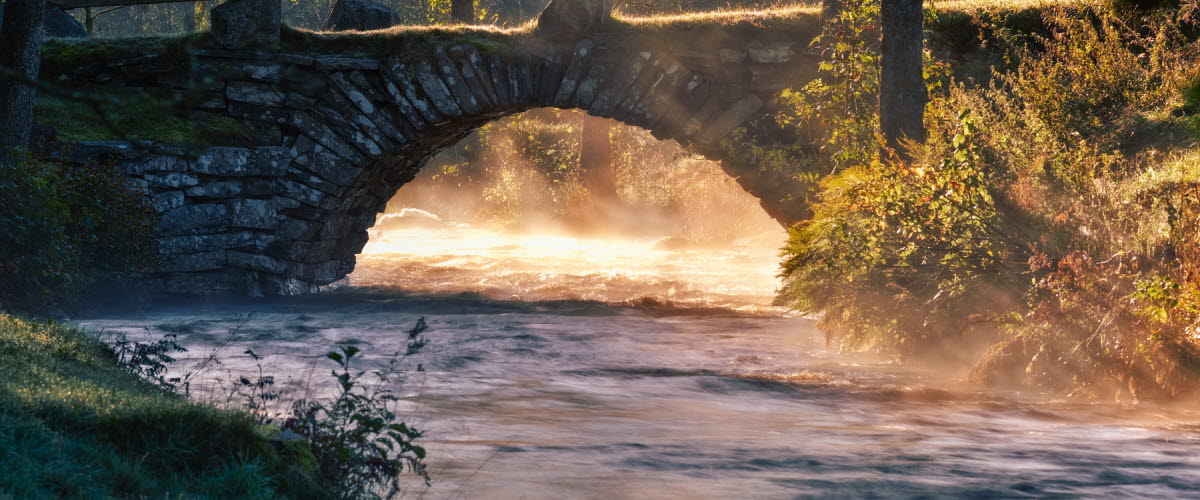 En bild på en gammal stenbro och en å. Det ligger en lätt dimma över vattenytan och solljuset är gyllene.