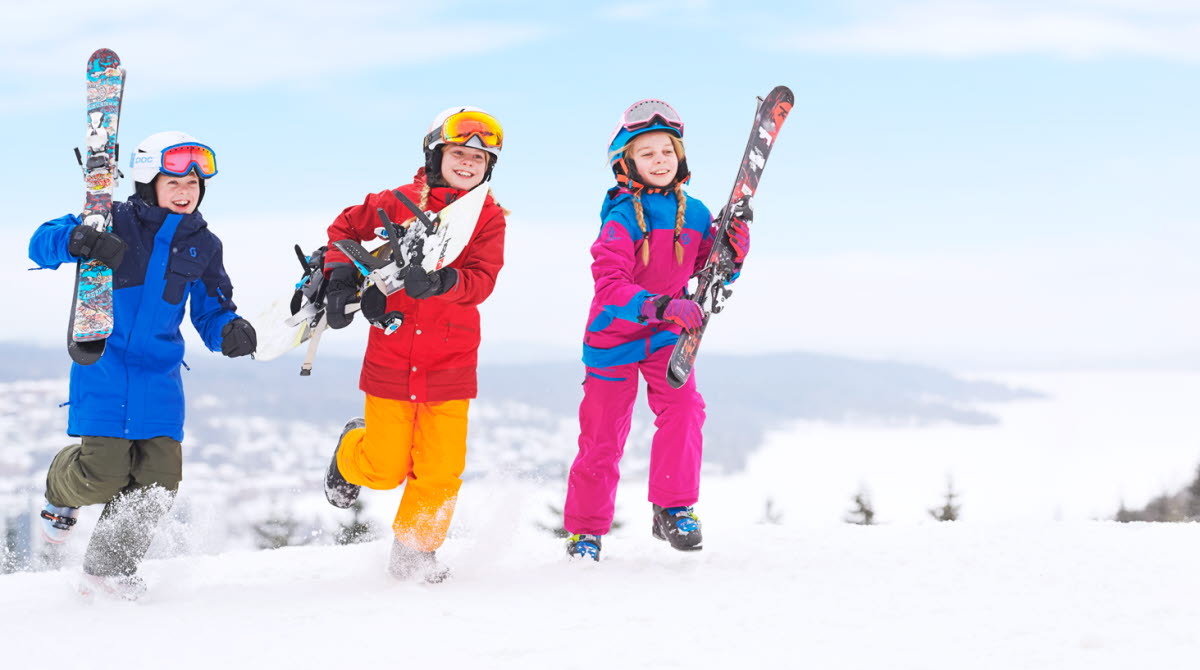 Three children in colorful clothes with skis in the arms running on top of Ulricehamn Ski Center.