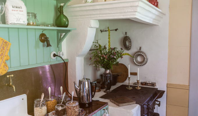 Ett gammaldags kök med vedspis och gröna träväggar. Frukostflingor står framdukat.