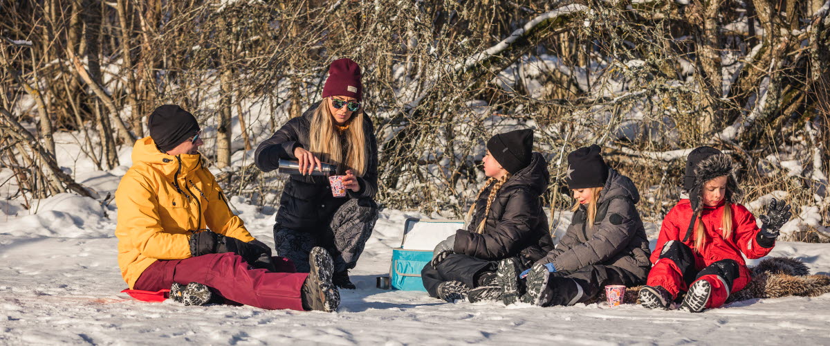 Man och kvinna och tre barn sitter ute i snön på filt och fikar.