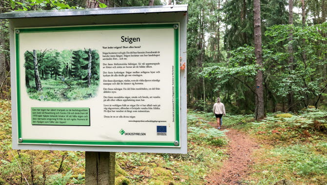 Informationsskylt längs Kulturstigen i Karleby. Längre fram ser man två personer som vandrar.