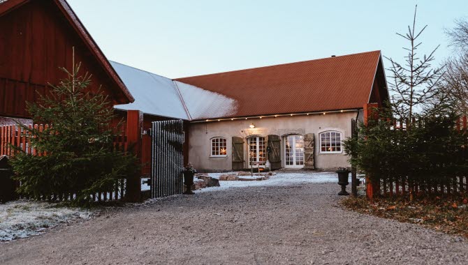 En gårdsbild med en gammal lada i vinkel. Framför entrén till gårdsplanen står två granar. Det är lite snö på marken.