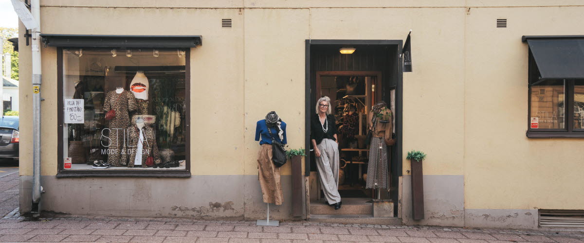 En kvinna står i en dörröppning i en butik som heter Stila i Hjo. Det är en byggnad i puts. Man ser även ett skyltfönster och utanför står en skyltdocka. 