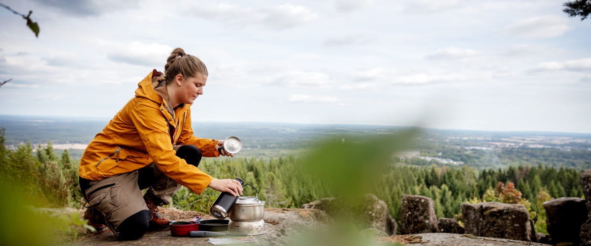Tjej i gul jacka sitter på kanten av ett berg och förbereder mat i trangiakök. Utsikten i bakgrunden är magisk. 