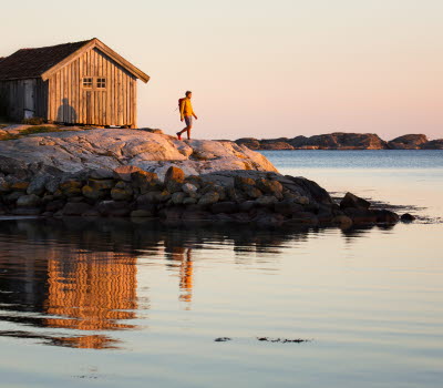En person införd en gul tröja står på en klippa framför en gammal, grå sjöbod på Sydkoster. Havet ligger blankt och solen är på väg att gå ned. 