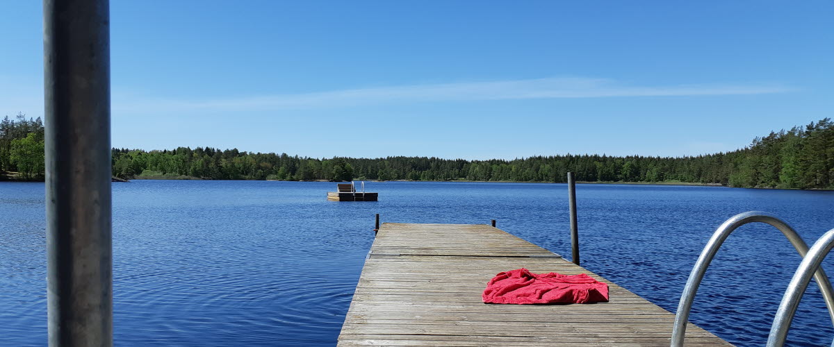 En brygga sticker ut i en blå sjö. En röd handduk ligger längre fram på bryggan.