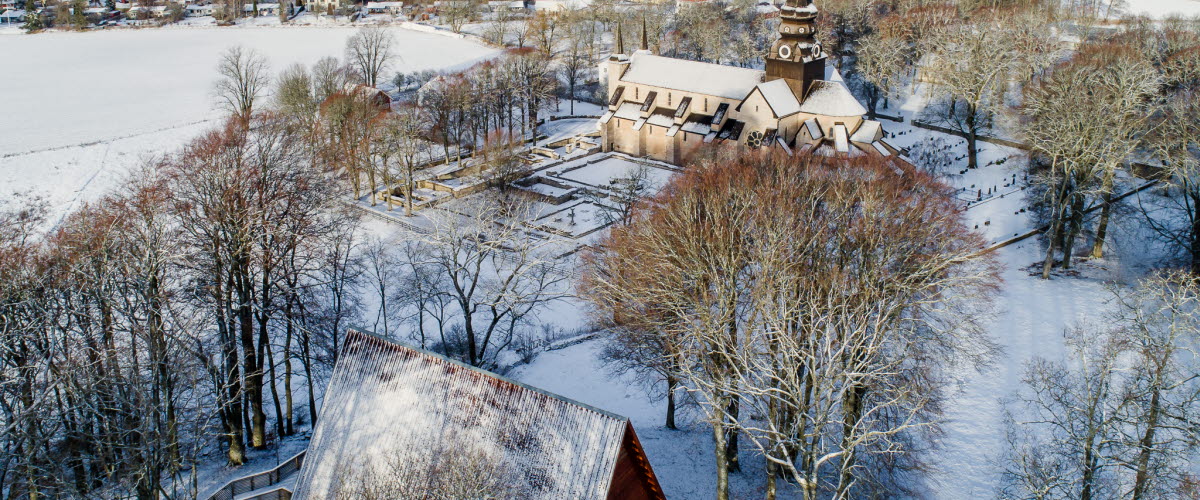 Ett drönarfoto av en stor stenkyrka och en träbyggnad. På marken ligger ett tunt lager snö.