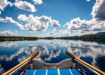 Bilden är tagen från en roddbåt och man skymtar en liten del av båten samt årorna, men mest fokus är det på den stilla sjön där himlen speglar sig.