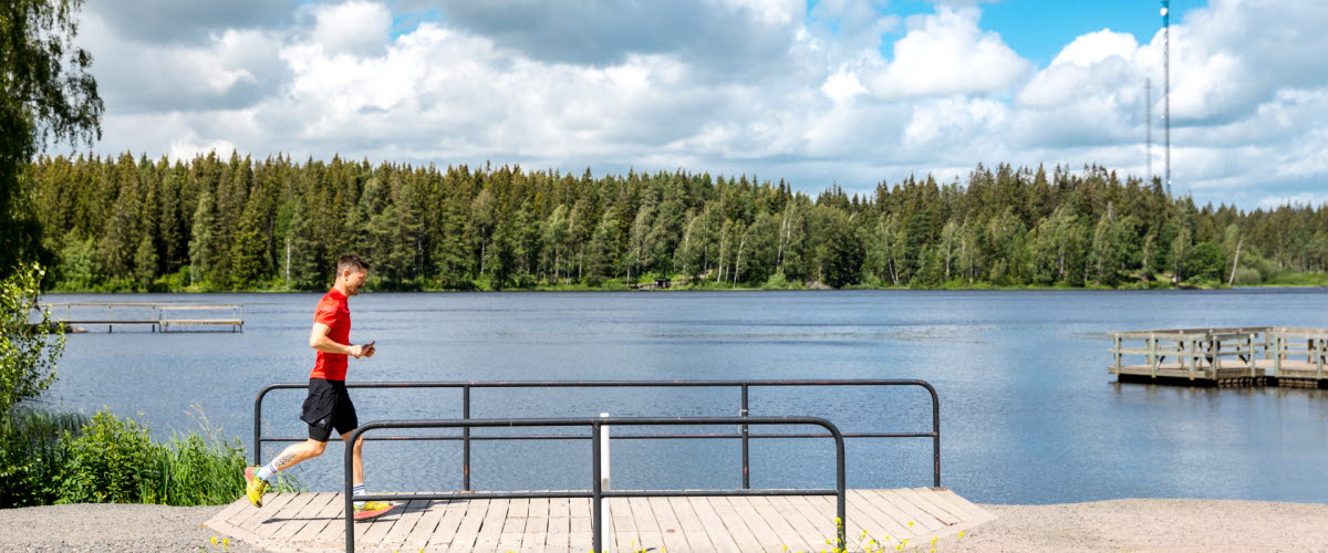 Man running along an exercise track, over a wide wooden bridge, behind the lake Hållsdammen spreads out and on the other side of it you can see the forest.