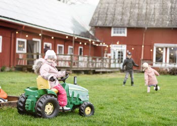 Ett barn sitter på en grön leksakstraktor framför en röd lada. Två andra barn leker i bakgrunden.