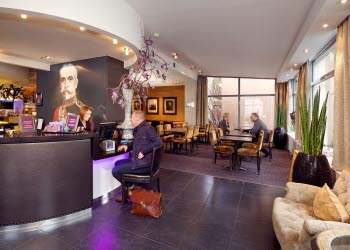 En man sitter på en stol i hotellreception och får hjälp av personal att checka in. I backgrunden syns del av matsalen där två personer sitter och samtalar.