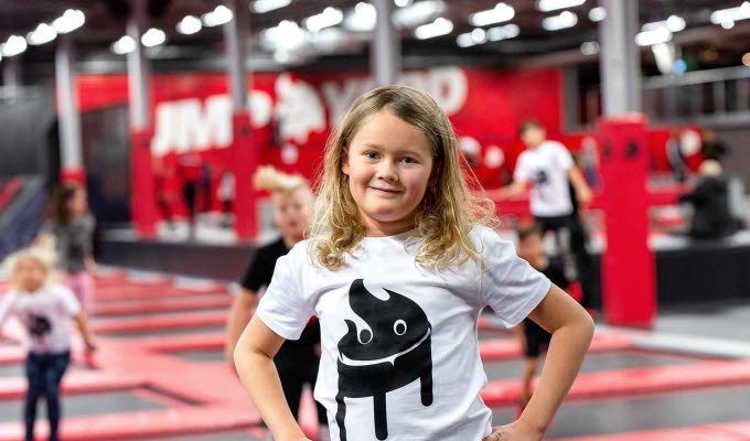 En flicka med blont hår och vit tröja tittar in i kameran. I bakgrunden ser man flera trampoliner från en trampolinpark breda ut sig. 