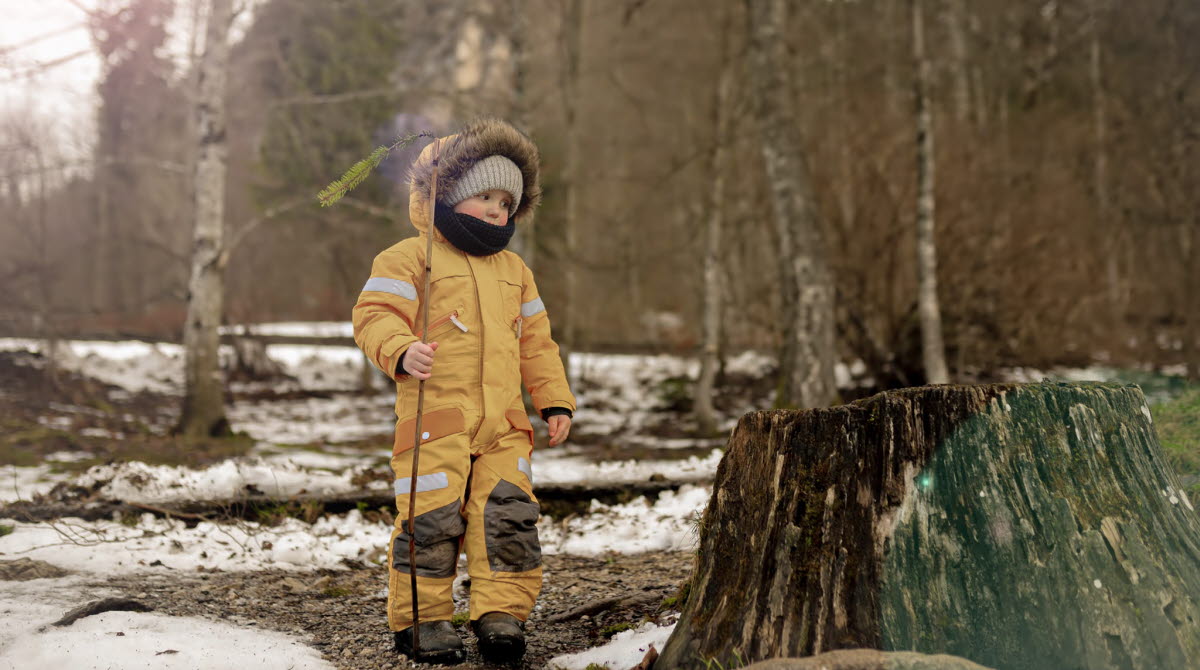 En bild på ett barn som leker i skogen. På marken ligger rester av snön kvar. Solen skiner.