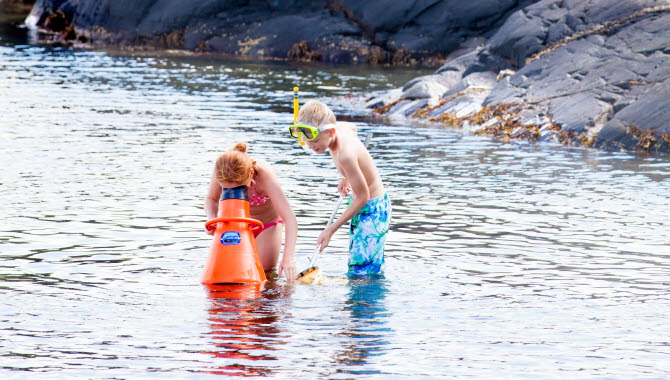 Två barn står i vattnet vid en strand. Ett av barnen har en stor, orange undervattenkikare och tittar ned på strandbottnen. Det andra barnet har en fiskehov.