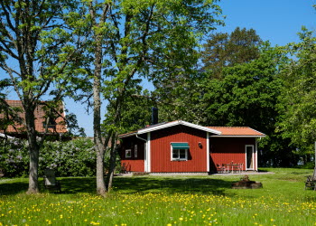 En röd stuga och runt om syns grönt gräs och gröna träd.