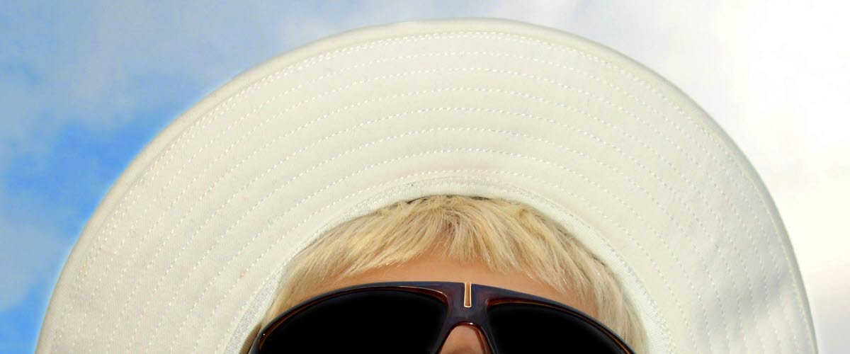 
A boy's face taken from below with blue sky and veil clouds in the background. The boy has black sunglasses and a white fabric hat