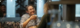 Two women are sitting at a table having coffee. One of them is smiling and holding a glass of coffee in her hand.