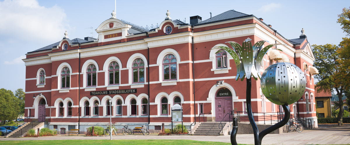 Tidaholms stadsbibliotek, en rosa byggnad med vita detaljer med ett konstverk av metall i en form av en blomma och en större boll