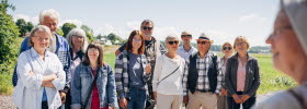 A group of people are standing and listening to a guide. In the background, you can see a blue sky and green landscape