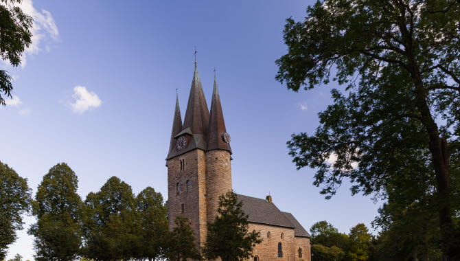 Gammal kyrka med tre torn. Framför kyrkan är det en kyrkogård med gravstenar och träd.