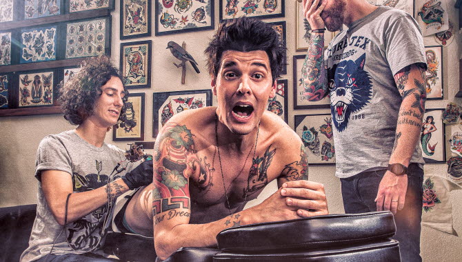 En man ligger på en tatueringsbänk och blir tatuerad. En person står bredvid men handen för ögonen.