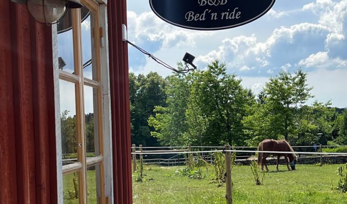 En närbild på en svart plåtskylt som hänger på en rödmålad husvägg. På skylten står det "Gyltegårdens B&B Bed' n ride"