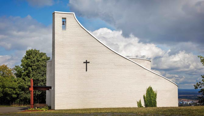 En vit modern stenkyrka. I bakgrunden ser man en milsvid utsikt över Skövde. Himlen är bitvis blå med dramatiska gråa moln. På kyrkan sitter ett litet svart kors.