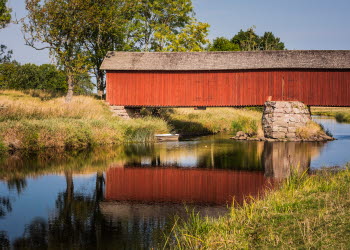 En långsmal bro som är inbyggd i falurött trä och har ett grått tak går över ett stilla vattendrag. Bron speglas av i vattnet.