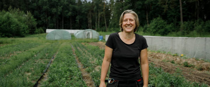 Susanne står framför sin odling i en glänta i skogen. Rader av grödor och tre växthus syns bakom henne.