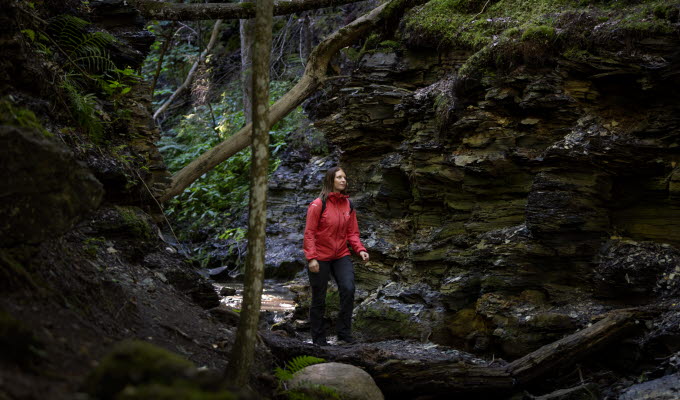 Kvinna i röd jacka och brunt hår går mellan stora stenblock där man ser bergslagren tydligt. 