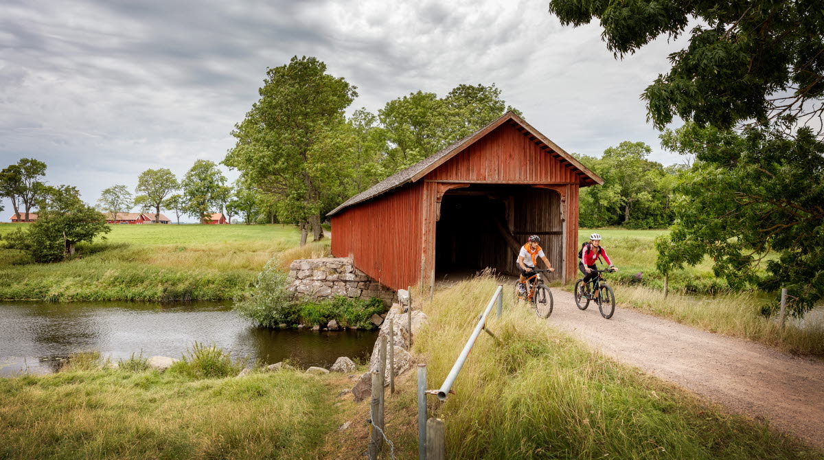 A man and a woman are cycling on a gravel road by the unique long bridge house, Vaholm's bridge house.