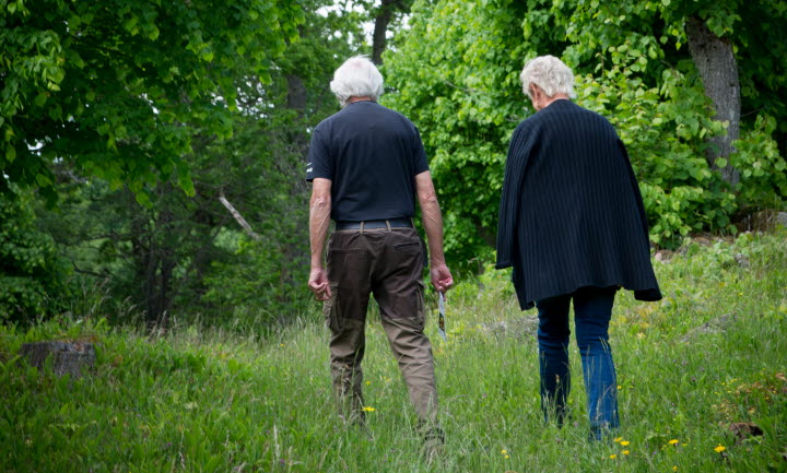 Ett äldre par i mörkblå kläder promenerar över en blomsteräng mot en skog.