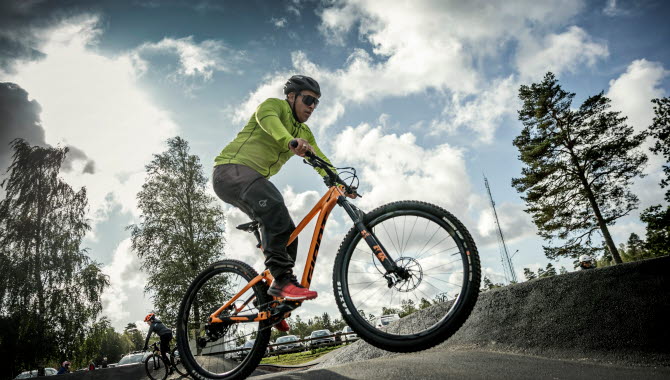 Kille i neonfärgad tröja och svarta byxor hoppar fram med sin orange mountainbike på den asfalterade pumptrackbanan.