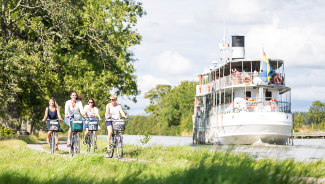 Fyra kvinnor cyklar i de gröna lummiga omgivningarna längs Göta Kanal. Bredvid dem i kanalen kör en passagerarångbåt.