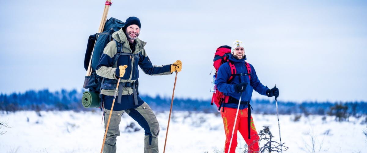 Två män står mitt i ett vinterlandskap i full vandrarutrustning med snöskor på fötterna. 
