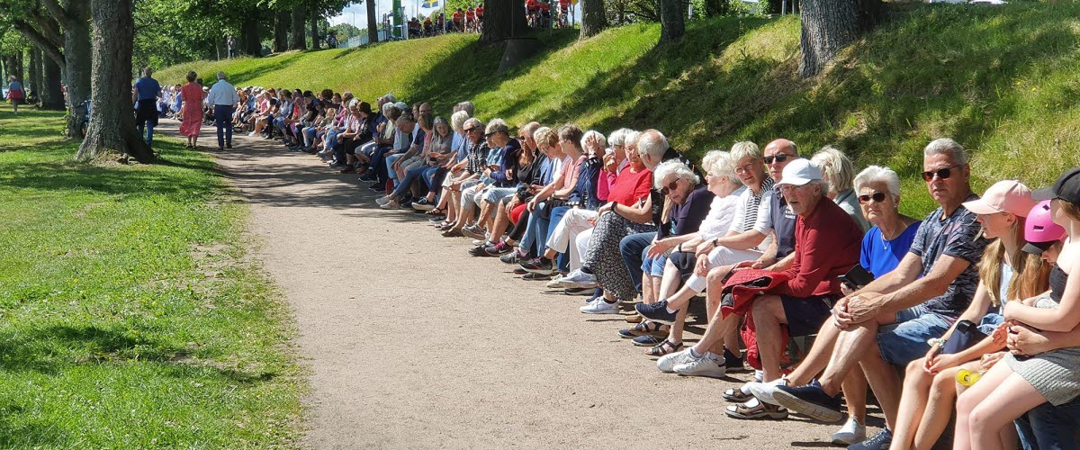 Människor sitter på Sveriges längsta bänk i Hjo, på Strandpromenaden en solig sommardag.