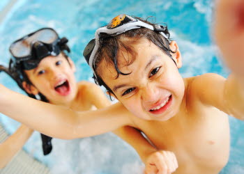 Två barn med simglasögon på huvudet busar i en bassäng.