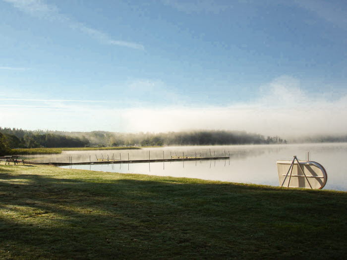 En bild på en sjö med dimma över vattenytan. En träbrygga leder ut till vattnet. I bakgrunden skymtar man skog. Framför sjön ligger en gräsmatta.