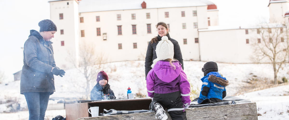 Vuxna och barn vid en grillplats med Läckö Slott i bakgrunden. På marken ligger snö.