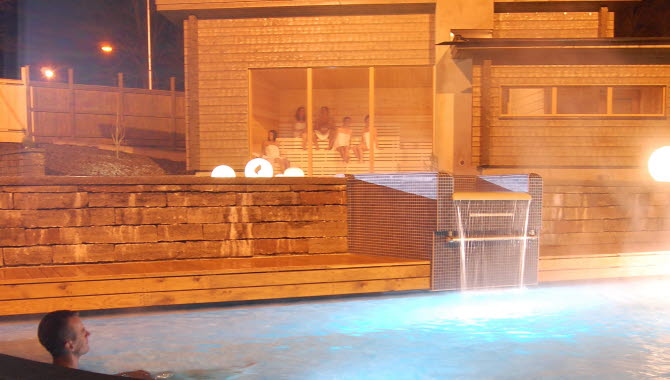 En man sitter i en ångande utomhuspool. Det är mörkt. I bakgrunden syns en träbastu med stora fönster.