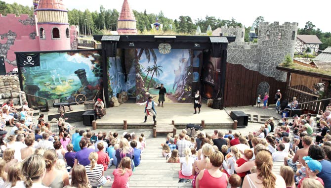 Piratshow på Daftölands stora scen. Läktaren är fylld av barn och familjer och i bakgrunden syns tornen av nöjesparkens lekslott.