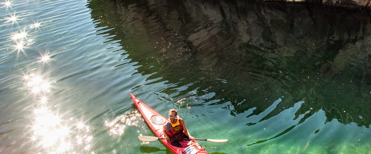A red kayak is paddeling past a cliff. It's a sunny day and the water is clear. 