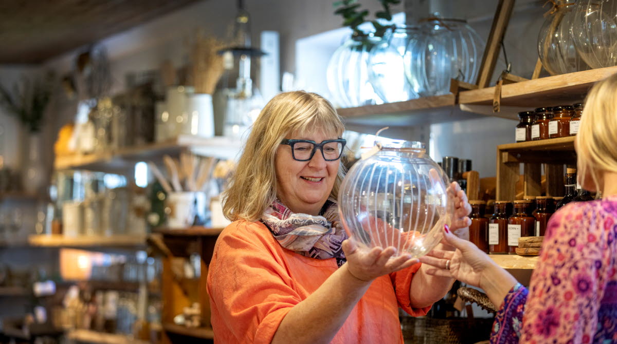 En kvinna i svarta glasögon och korallfärgad tröja står och tittar på en glasvas i en butik. 
