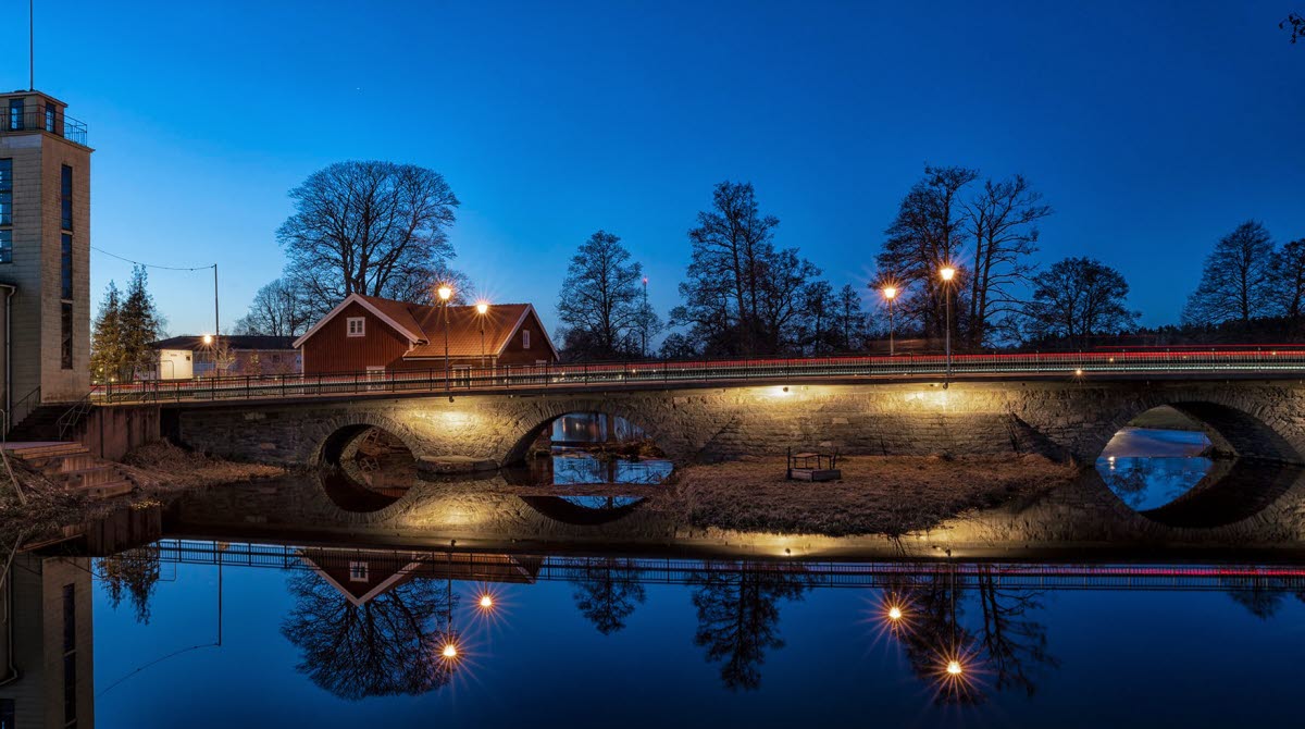an evening picture in front of a stone bridge reflecting in the water. A red house and a gray house on the side of the bridge.