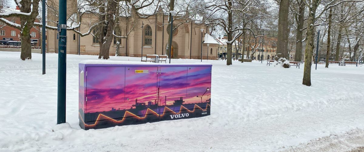 En vintrig bild från kyrkparken med kyrkan i bakgrunden där ett elskåp som klätts med en bild på Volvofabriken i skymning syns.