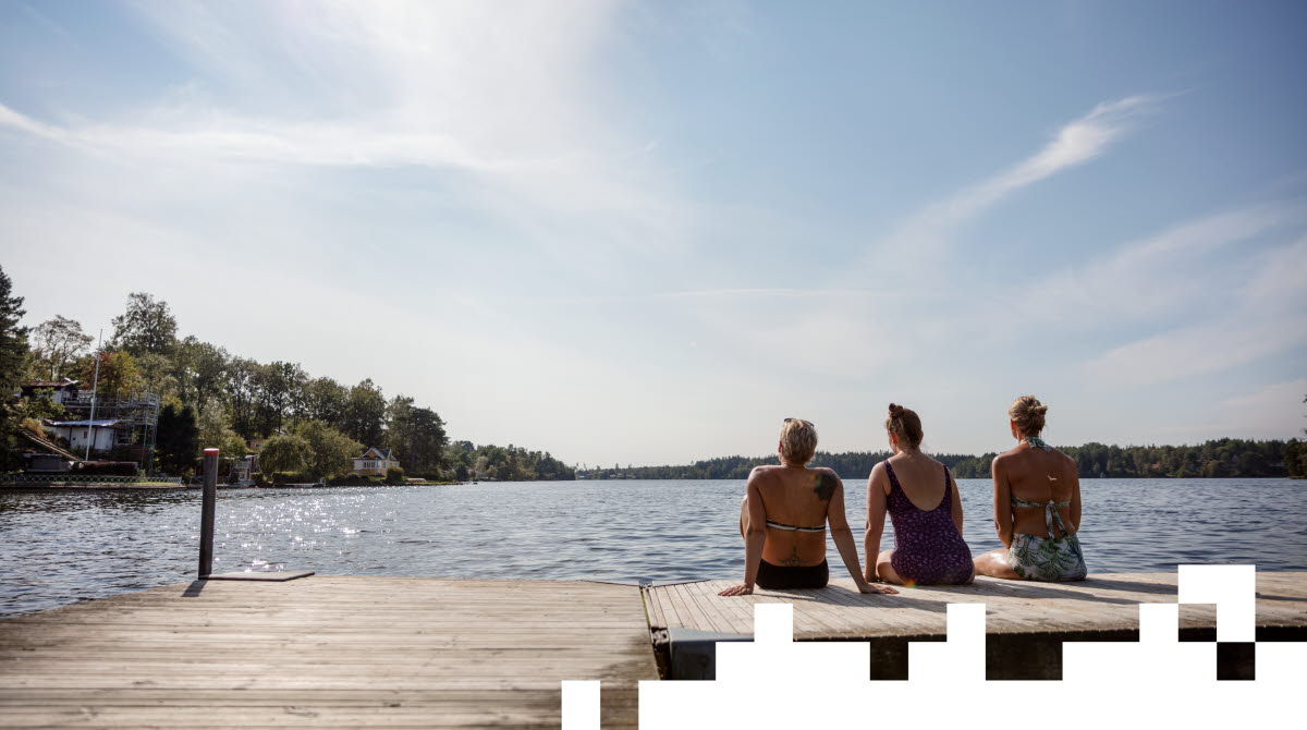 Three women in swimwear sit with their backs to the camera on a bridge and look out over a lake.